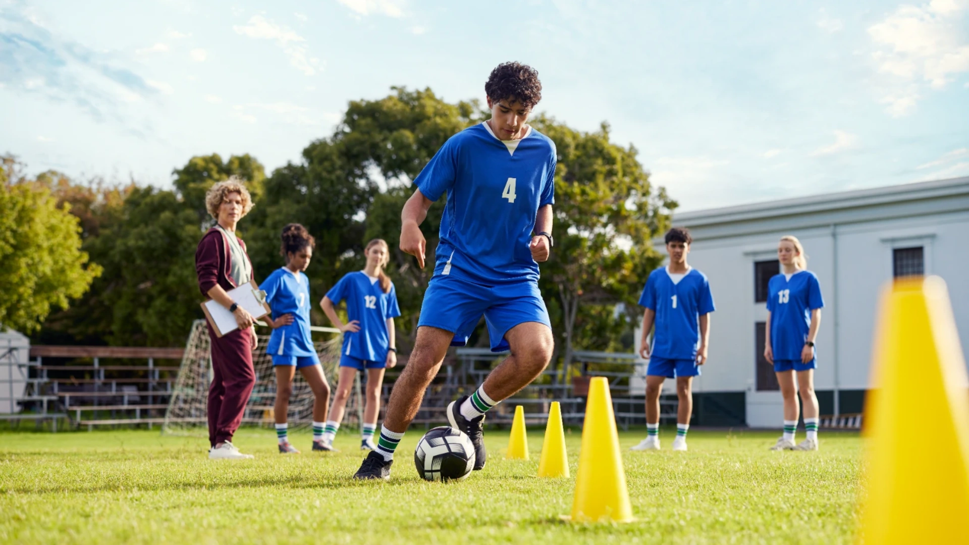 Young footballers in blue kit running a cone drill at sports club training while their coach watches